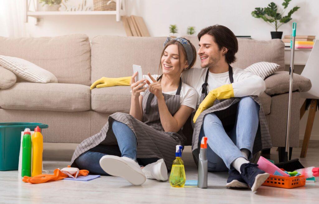 Junges Paar mit Reinigungshandschuhen sitzt lachend vor Sofa und nutzt Smartphone im modernen Wohnzimmer.